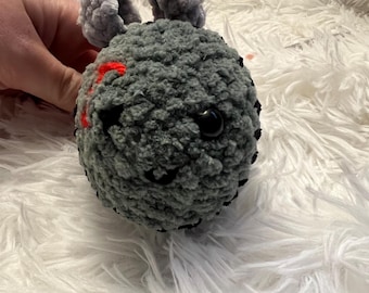 May include: A handmade crochet creature with a gray body, black stitching, and red accents. It has two gray, fluffy ears and a black eye. The plush toy is held against a white, fluffy background.