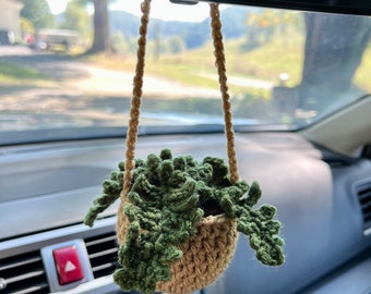 May include: A handmade crochet car air freshener featuring a small, woven basket holding a green, leafy plant. The basket is suspended by a tan rope, adding a touch of nature to the car's interior. The plant is a vibrant green.