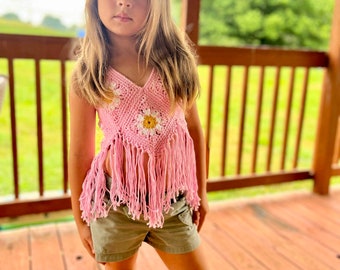 May include: A pink crochet top with a V-neck and fringe detail. The top features a daisy design and is paired with khaki shorts. The outfit is suitable for children's wear.