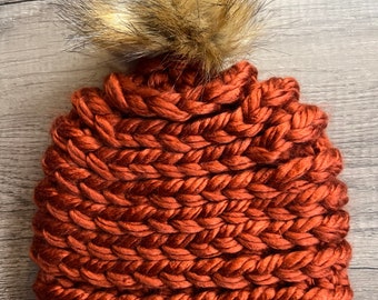 May include: A chunky knit beanie in a warm rust color, topped with a fluffy, light brown faux fur pom-pom. The hat's texture is clearly defined, showcasing the thick yarn used in its construction. This cozy winter hat is perfect for cold weather.