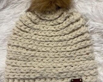 May include: Cream-colored knit beanie with a large, fluffy brown pom-pom on top. The hat features a chunky knit design and a small, brown leather tag with gold rivets. The hat is displayed on a white, fluffy surface.