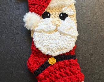 May include: Crocheted Christmas stocking shaped like Santa Claus. The stocking features a red hat and suit, a cream-colored face and beard, and black eyes. A gold button accents the suit. The toe is black and white.