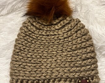 May include: A tan-colored, hand-knitted beanie with a large, fluffy brown pom-pom on top. The hat features a thick, textured stitch and a small, brown leather tag with text. The beanie is displayed on a white, fluffy surface.