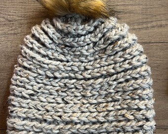 May include: A gray knit beanie with a large, fluffy, brown faux fur pom-pom on top. The hat is made of thick, chunky yarn, and the texture is clearly visible. The beanie is a neutral color, suitable for various outfits.