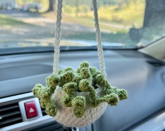 May include: A white crocheted hanging planter with a green succulent plant inside, suspended from a car's rearview mirror. The planter is round and has a textured appearance, adding a touch of handmade charm to the car's interior.