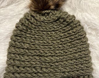 May include: A handmade olive green knit beanie with a fluffy brown faux fur pom-pom on top. The hat has a ribbed texture and a small burgundy tag on the side. The beanie is displayed on a white, fluffy surface.