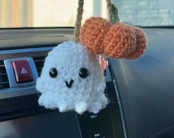 May include: A handmade white crochet ghost car charm with black eyes and a smiling mouth. Above the ghost are two orange crochet cherries with a brown string and green leaf. The charm is hanging in a car.