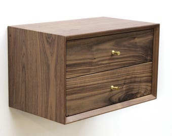 Pode incluir: Uma mesa de cabeceira de madeira castanho escuro com duas gavetas e puxadores de latão. A mesa de cabeceira está montada na parede.