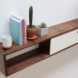 May include: A wall-mounted walnut wood shelf with two white drawers and a lower open shelf. The shelf has a cactus, a succulent, a pitcher, and a stack of books on it.