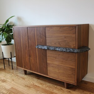 May include: A mid-century modern style wooden cabinet with four doors and a drawer. The cabinet is made of dark brown wood and features a unique, textured, dark gray accent piece. The cabinet is set against a white wall.