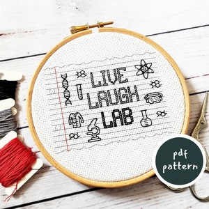 May include: A white cross-stitch pattern with the text "Live Laugh Lab" in black. The pattern is surrounded by small science-themed icons, including a DNA strand, a beaker, a test tube, and a microscope. The pattern is framed in a wooden hoop.