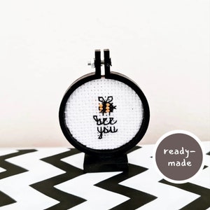 May include: A small, round embroidery hoop with a white fabric background. The fabric has a black and yellow bee embroidered on it with the text "bee you".
