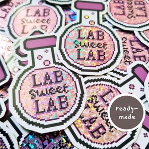 May include: A selection of stickers with a lab flask design. The stickers display the text "LAB Sweet LAB" in a pixelated font, with a pink and glittery background. The flask outline is black and the top is purple.