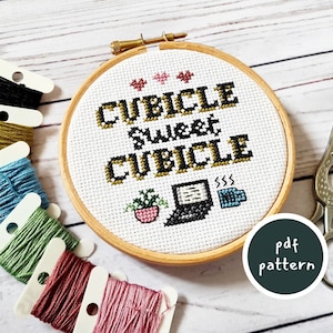 May include: A cross-stitch embroidery hoop with a white fabric background and the text "Cubicle Sweet Cubicle" in black and gold lettering. The design also includes a potted plant, a laptop, and a coffee cup.