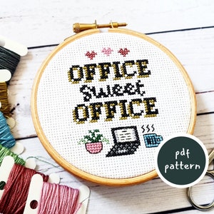 May include: A white embroidery hoop with the text "Office Sweet Office" in black and gold lettering. There are three small pink hearts at the top of the hoop. A small potted plant, a laptop, and a coffee cup are embroidered below the text. The hoop is surrounded by colourful embroidery floss.