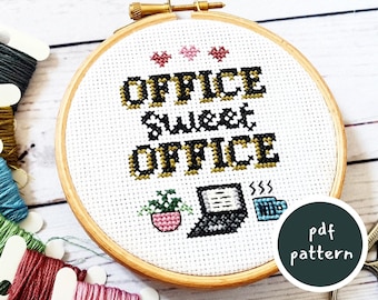 May include: A white embroidery hoop with the text "Office Sweet Office" in black and gold lettering. There are three small pink hearts at the top of the hoop. A small potted plant, a laptop, and a coffee cup are embroidered below the text. The hoop is surrounded by colorful embroidery floss.