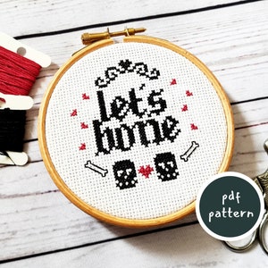 May include: A cross-stitch embroidery hoop featuring the phrase "let's bone" in black, with skull and bone motifs. The design is on white fabric, framed in a wooden hoop. Red and black thread spools are visible.