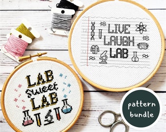 May include: Two embroidery hoops with cross-stitch patterns. The top hoop has a white background with black text that reads "Live, Laugh, Lab" and includes science-themed icons. The bottom hoop has a white background with black text that reads "Lab Sweet Lab" and includes colorful beakers. The hoops are on a white wooden surface with embroidery floss and scissors.
