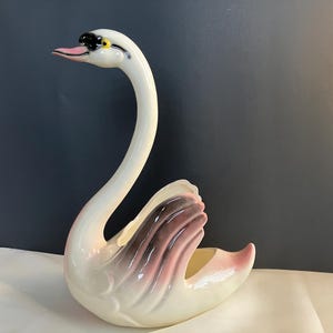 May include: A ceramic swan figurine with a long, curved neck and a graceful pose. The swan is primarily white with pink and brown accents on the wings. The beak is pink, and the eyes are black and yellow. This decorative piece is likely a planter or vase.