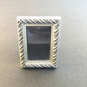 May include: A small, rectangular silver picture frame with a decorative rope-like border. The frame has a dark interior and is designed to hold a small photograph or artwork. The frame is displayed on a dark surface.