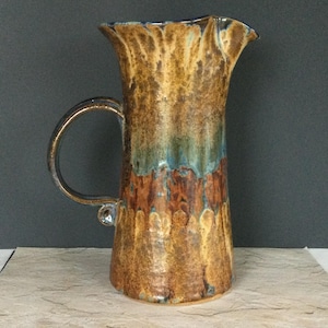 May include: A tall, handcrafted ceramic pitcher with a wide, ruffled rim and a curved handle. The pitcher features a textured surface with a blend of brown, gold, and teal colors, creating a rustic, earthy aesthetic.
