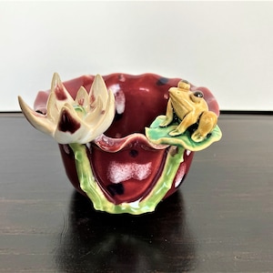 May include: A small ceramic bowl with a red and white speckled glaze. The bowl is shaped like a lotus flower with a green stem and leaves. A small yellow frog sits on a green leaf on the rim of the bowl.