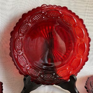 May include: A round, translucent ruby red glass plate featuring a scalloped edge and a decorative border with embossed diamond and oval patterns. The center of the plate displays a sunburst design radiating from the middle toward the rim.