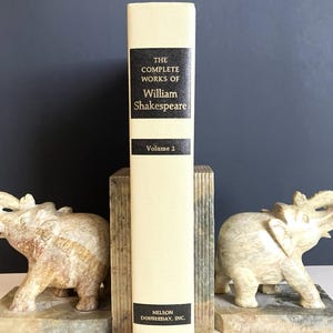 May include: A cream-colored book titled "The Complete Works of William Shakespeare, Volume 2" stands between two carved stone elephant bookends. The bookends and book are on a gray surface with a dark blue background.