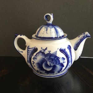 May include: A white porcelain teapot with a blue and white floral design. The teapot has a lid with a knob on top. The teapot is sitting on a dark surface.