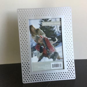 May include: A silver metal photo frame with a dotted pattern. The frame has a white mat and a clear plastic insert. The frame is designed to hold a 4x6 photo. The text "4x6 Photo Frame" is printed on the mat.