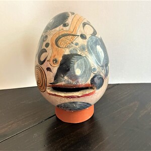 May include: A ceramic egg-shaped piggy bank with a painted design of birds, leaves, and swirls in shades of blue, yellow, and white. The piggy bank has a slot for coins on the side.