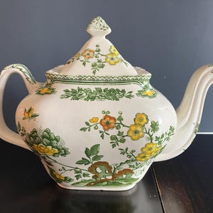May include: A cream-colored ceramic teapot featuring a decorative pattern of green leaves and yellow and orange flowers. The teapot has a matching lid with a pointed finial, a curved handle, and a spout. The floral design covers the body and lid.