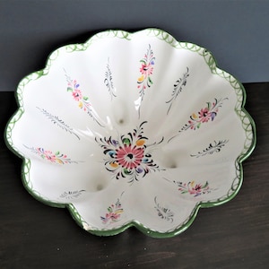 May include: A white ceramic bowl with a scalloped edge and green trim. The bowl is decorated with hand-painted flowers in shades of pink, red, yellow, and green.