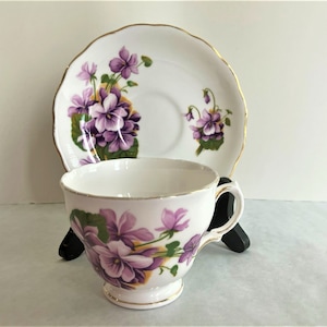 May include: A white teacup and saucer with a gold trim and purple violet floral pattern.