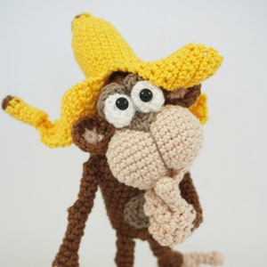 Könnte beinhalten: Ein gehäkelter Affe, der einen gelben Bananenhut trägt. Der Affe hat braunes Fell und ein rosafarbenes Gesicht. Der Affe schaut nach rechts und hat seine Hand am Kinn.