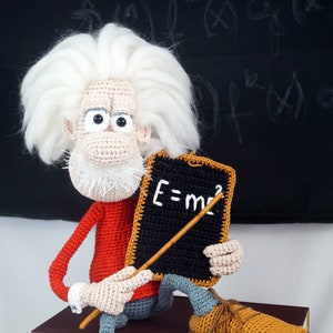 Puede incluir: Mu&ntilde;eco de Einstein tejido a ganchillo con pelo y barba blancos, con un su&eacute;ter rojo, pantalones azules y zapatos marrones. Sostiene una peque&ntilde;a pizarra con la ecuaci&oacute;n E=mc&sup2;. El mu&ntilde;eco est&aacute; sentado sobre una pila de libros.