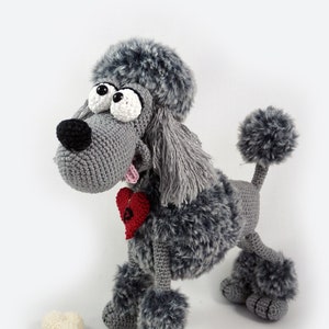 May include: A grey crocheted poodle toy with a red heart and a bone. The poodle has a fluffy tail and large, expressive eyes.