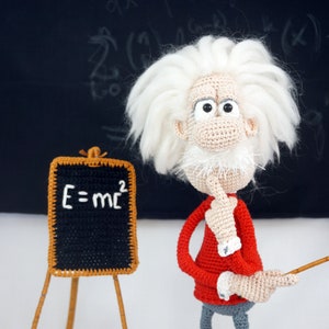 May include: A crocheted doll of a scientist with wild white hair, a red long-sleeved shirt, and gray trousers, standing next to a small easel. The easel holds a black board with the white text E=mc2. The doll wears brown shoes and holds a pointer.