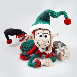 May include: A green and red crocheted elf doll with a white pom-pom hat, lying on its side with its head resting on its hand. The elf is smiling and has large eyes. The elf is next to a small gift box with a silver ribbon.