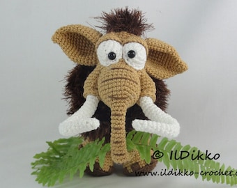 May include: A crocheted brown and white woolly mammoth toy with large eyes and a fern plant in front of it. The toy has a brown and white striped trunk and white crocheted tusks. The toy is sitting on a white surface. © Ildikko www.ildikko-crochet.com
