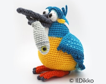 May include: A crocheted blue and yellow kingfisher holding a gray crocheted fish in its beak. The kingfisher has large, white eyes and a black beak. The fish has a green eye. The kingfisher is sitting on a white surface. © Ildikko www.ildikko-crochet.com