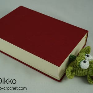 May include: A green crocheted dragon bookmark peeking out from the side of a red hardcover book. The bookmark has white eyes and a pink tongue.