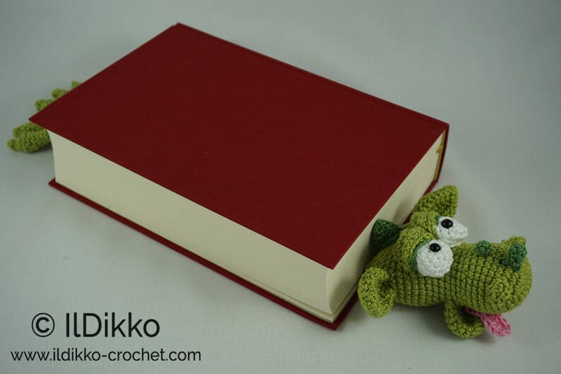 May include: A green crocheted dragon bookmark peeking out from the side of a red hardcover book. The bookmark has white eyes and a pink tongue.