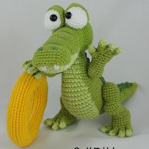 May include: A green crocheted crocodile toy with large white eyes and black pupils, holding a bright yellow crocheted ring. The text "&copy; ILDikko www.ildikko-crochet.com" is printed in black at the bottom right of the image.
