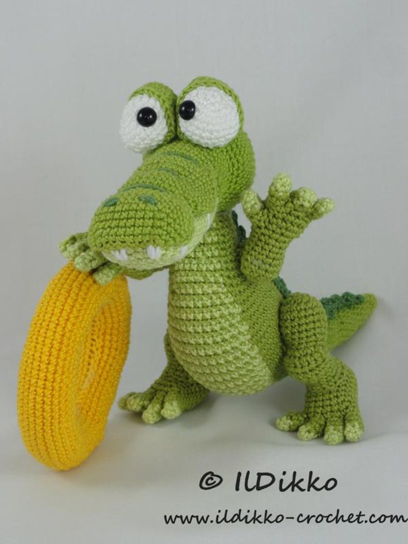 May include: A green crocheted crocodile toy with large white eyes and black pupils, holding a bright yellow crocheted ring. The text "&copy; ILDikko www.ildikko-crochet.com" is printed in black at the bottom right of the image.