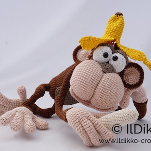 May include: A crocheted monkey toy wearing a yellow banana hat. The monkey is brown and pink with large black eyes. The monkey is holding a white crocheted banana in its hand. © IlDikko www.ildikko-crochet.com