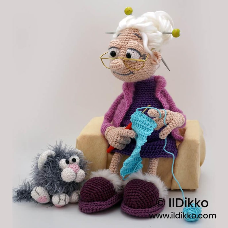 May include: A crocheted doll of a smiling elderly woman with white hair, glasses, and a purple outfit, knitting with blue yarn. A gray cat and purple slippers with white trim sit nearby. The doll is seated on a tan cushion.