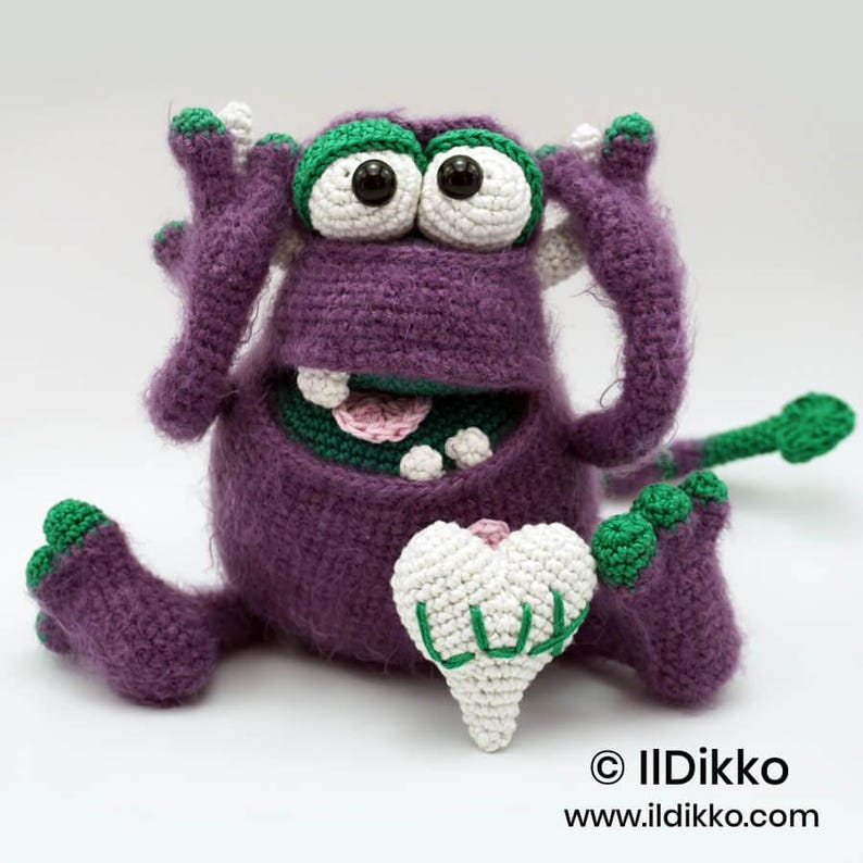 Puede incluir: Un juguete de monstruo tejido a ganchillo de color morado con detalles verdes en pies, manos y ojos. Tiene grandes ojos blancos con pupilas negras, una boca ancha con dientes blancos y sostiene un coraz&oacute;n tejido blanco. Texto: &copy; IlDikko www.ildikko.com.