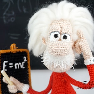 May include: A crocheted doll featuring a beige face, white hair, and a white beard, wearing a red long-sleeved shirt with white cuffs. The doll holds a small black chalkboard with the white text E=mc2 and a piece of white chalk in one hand.