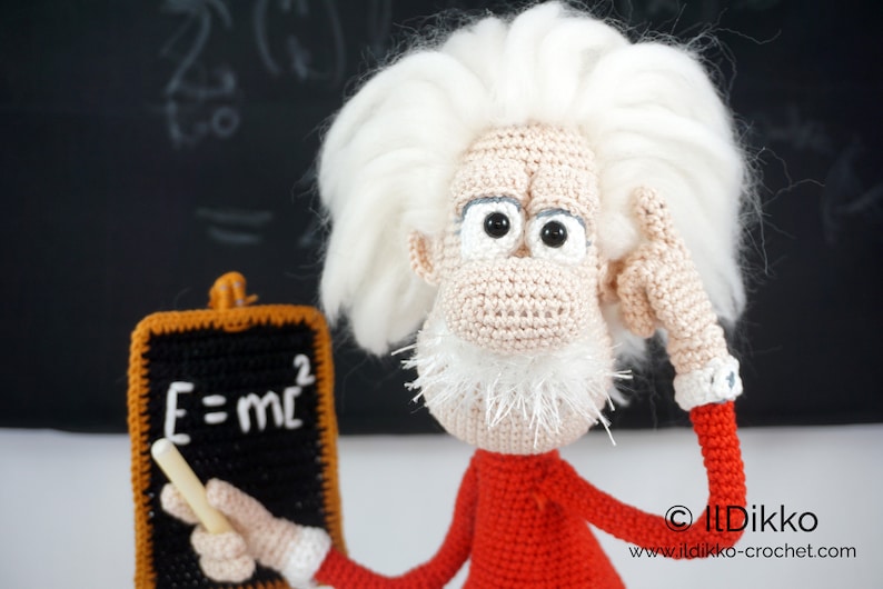 May include: A crocheted doll featuring a beige face, white hair, and a white beard, wearing a red long-sleeved shirt with white cuffs. The doll holds a small black chalkboard with the white text E=mc2 and a piece of white chalk in one hand.
